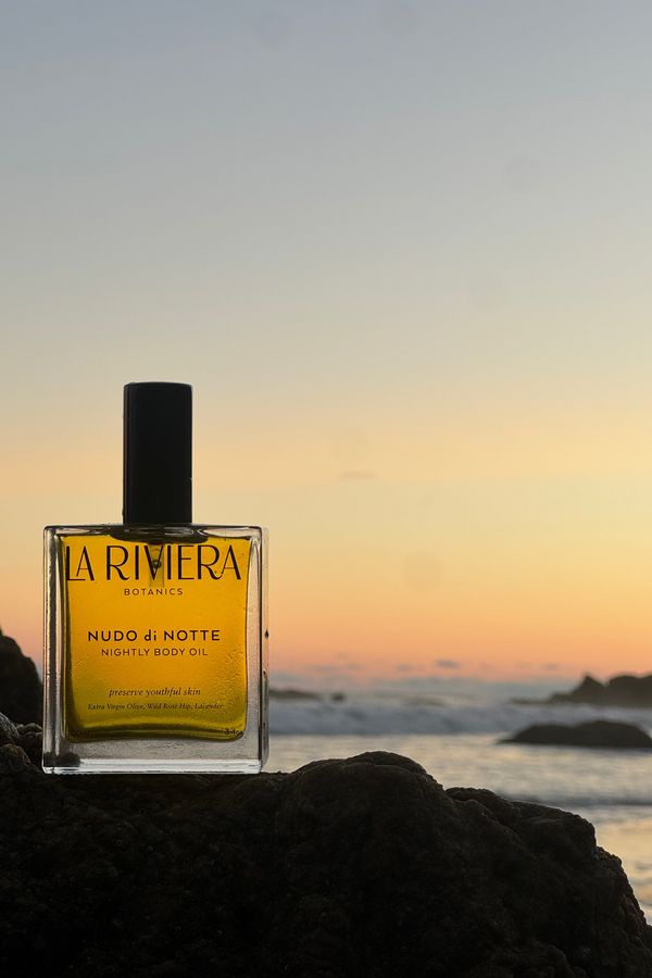 Bottle of La Riviera Nudo di Notte body oil on a rock with a sunset over the ocean.